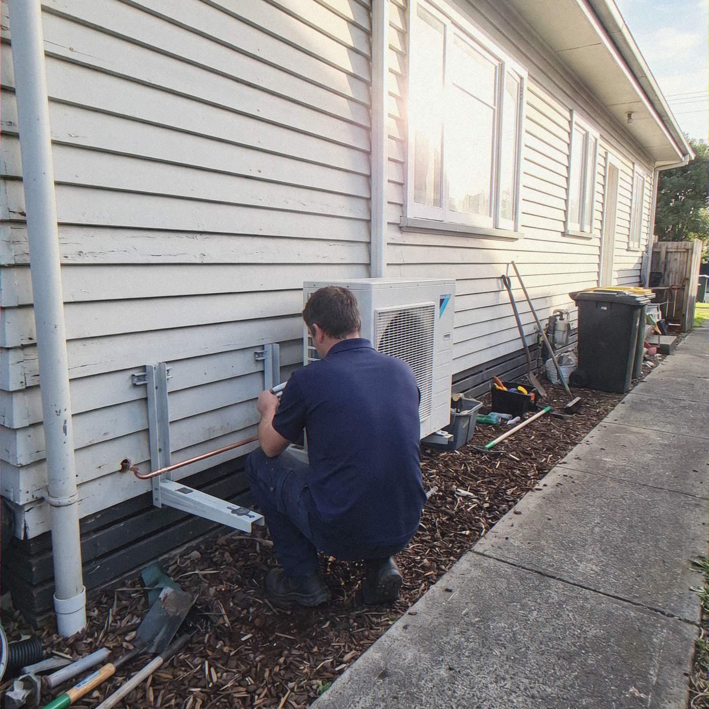 Installer connecting pipework to a heat pump outdoor unit on the side of a weatherboard house